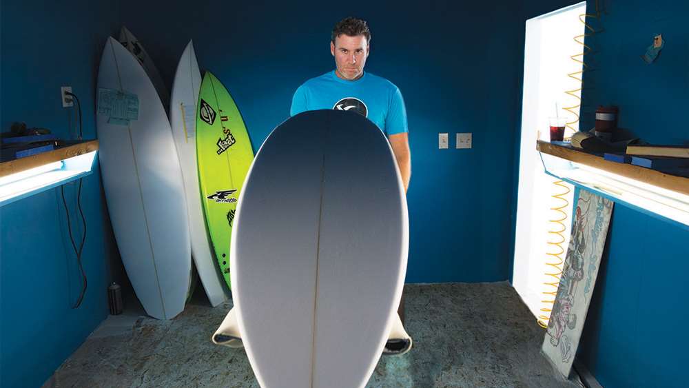 Man holding a surfboard in a workshop with blue walls.