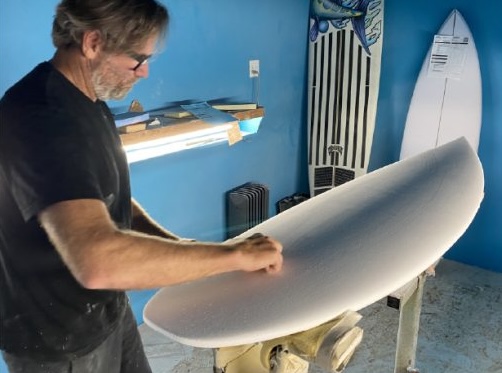 Man shaping a surfboard in a workshop with blue walls.