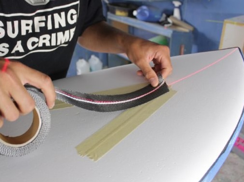 Person applying tape to a surfboard in a workshop.
