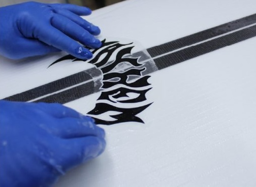 Hands in blue gloves applying a black design on a surfboard.