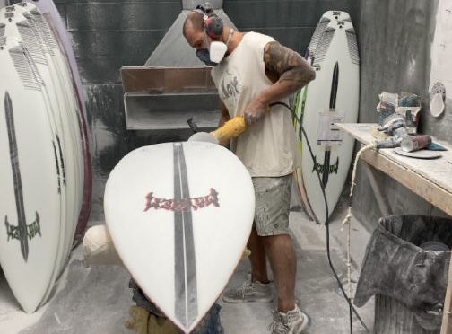 Person shaping a surfboard in a workshop with multiple boards.