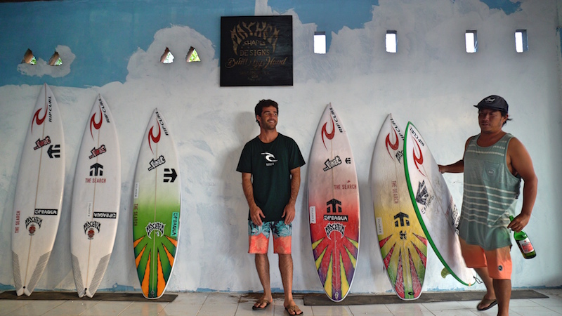Two surfers stand beside surfboards on a wall.