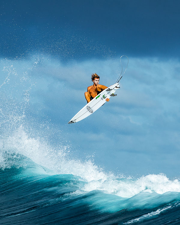 A surfer performs an aerial maneuver above a large wave, with water splashing around him. The sky is partly cloudy, creating a dynamic backdrop for the action.