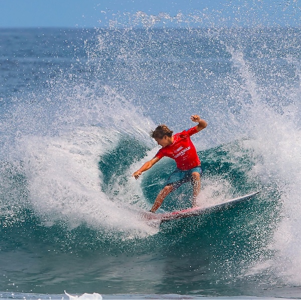 Child surfing on a wave, wearing a red shirt.