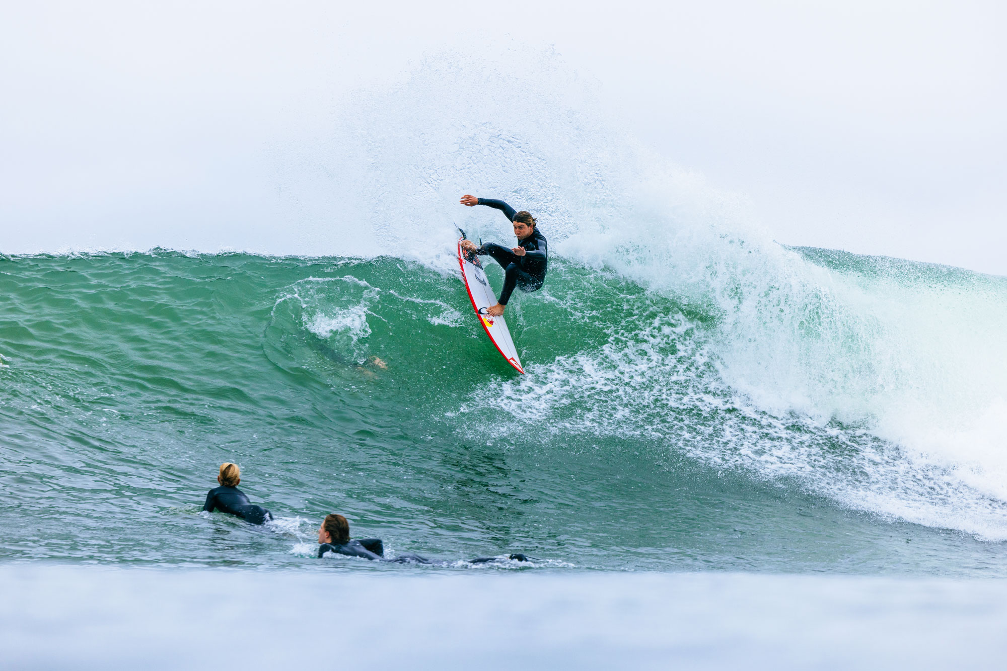 Surfer performing a maneuver on a large wave, two others paddling.
