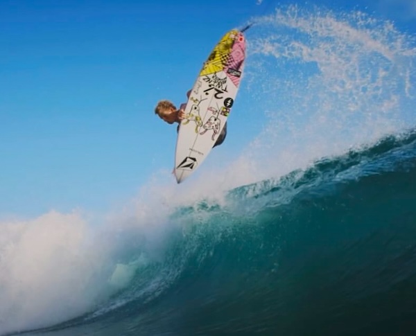 Surfer performing a trick above a wave against a blue sky.