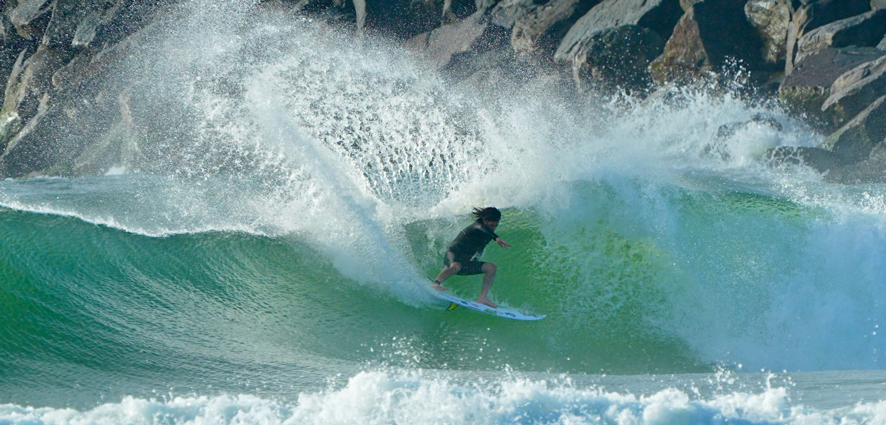 Surfer riding a large wave near rocky coastline.