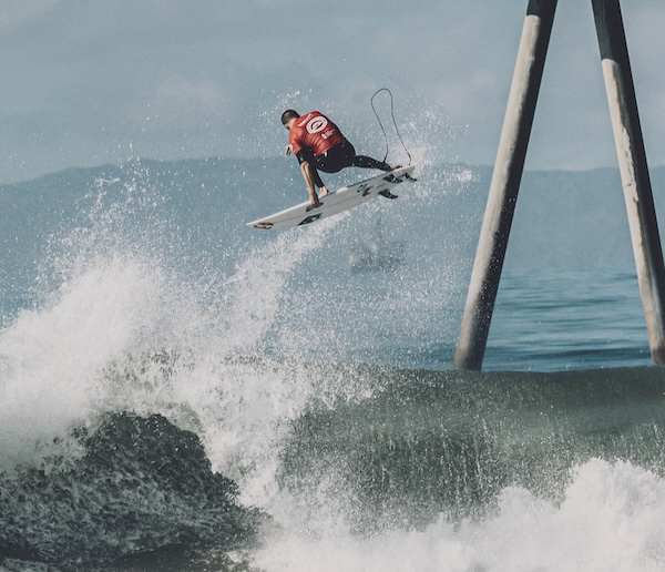 Surfer performing a jump over a wave near pilings.
