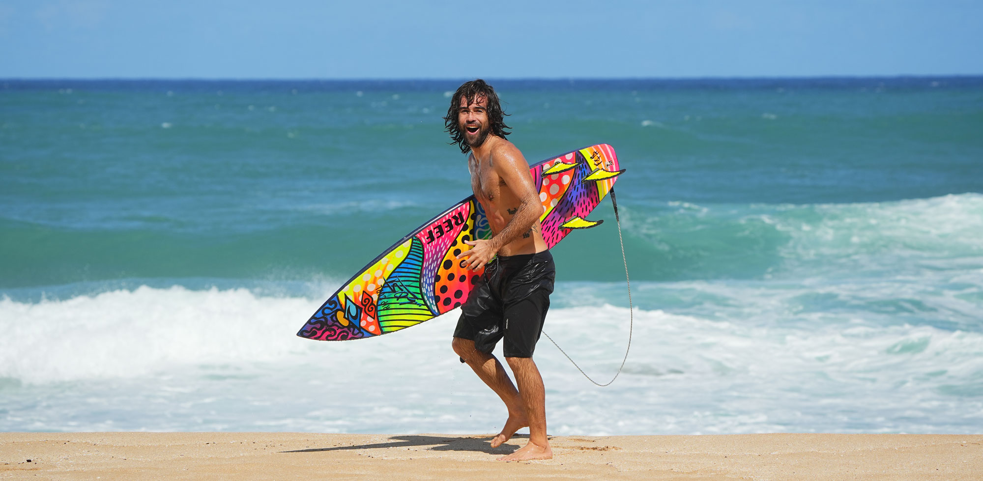 Surfer smiling while holding a colorful surfboard on the beach.
