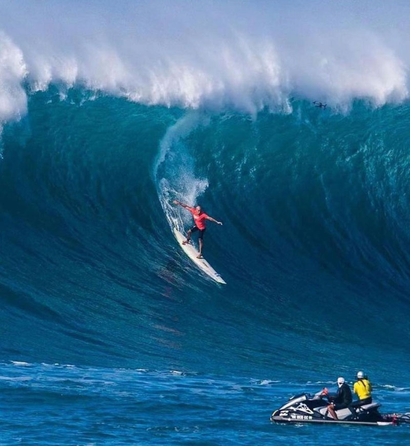 Surfer riding a large wave with jet ski nearby.