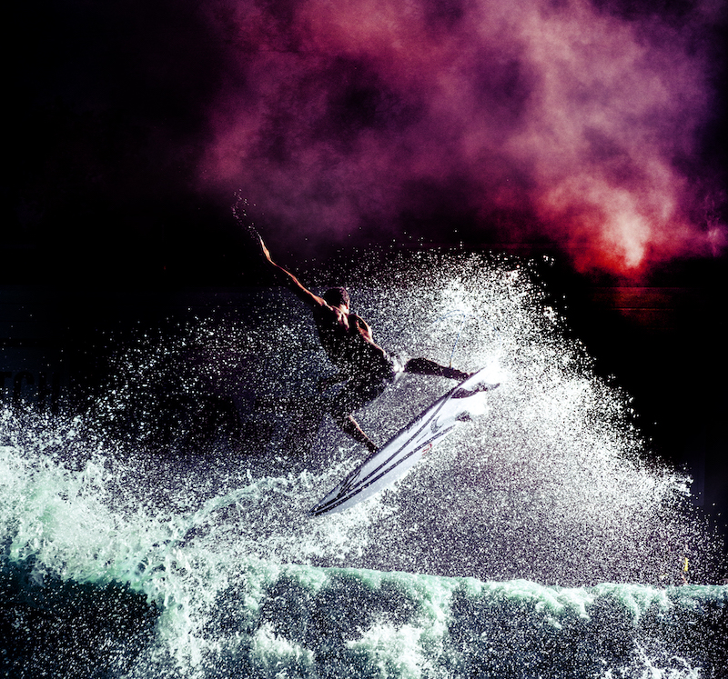 Surfer performing a trick above crashing waves with colorful background.