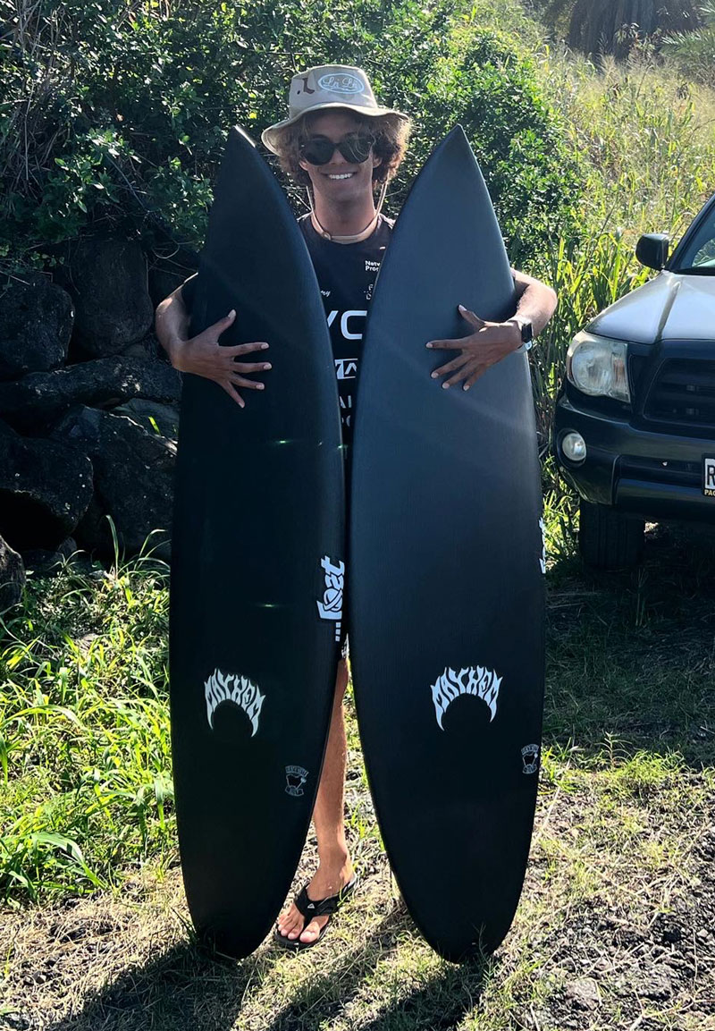 Person holding two black surfboards outdoors, wearing sunglasses and a hat.