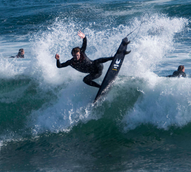 Surfer performing a maneuver on a wave, with others in background.