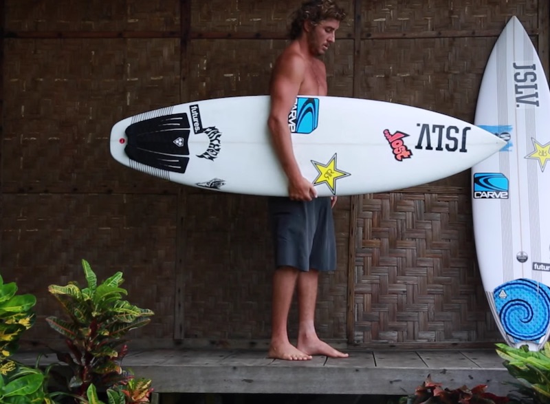 Shirtless man holding a surfboard on a wooden deck.