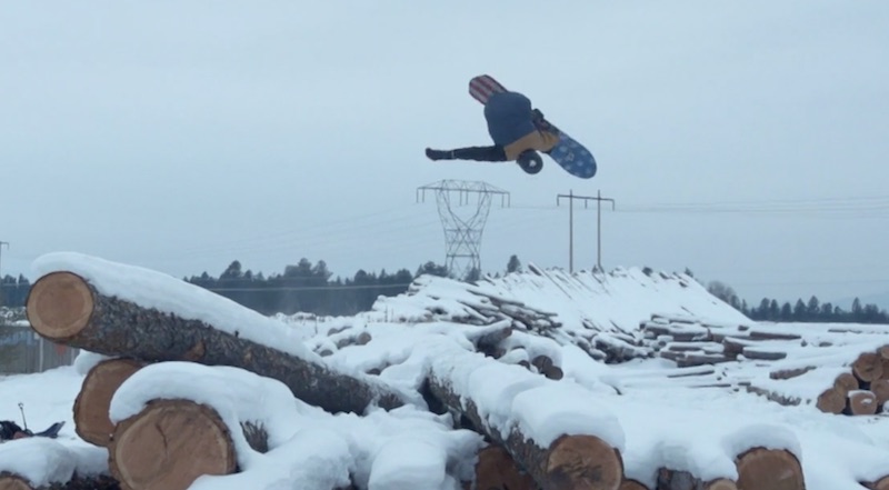 Snowboarder performing a jump over logs in a snowy landscape.
