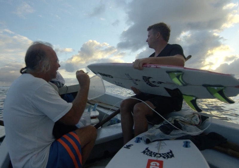 Two men on a boat with surfboards, ocean in background.