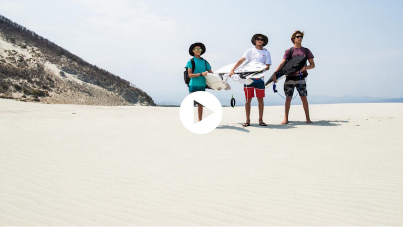Three surfers standing on a sandy beach with surfboards.