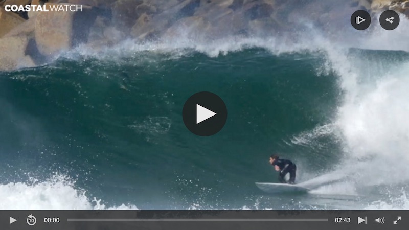 Surfer riding a large wave near rocky shoreline.