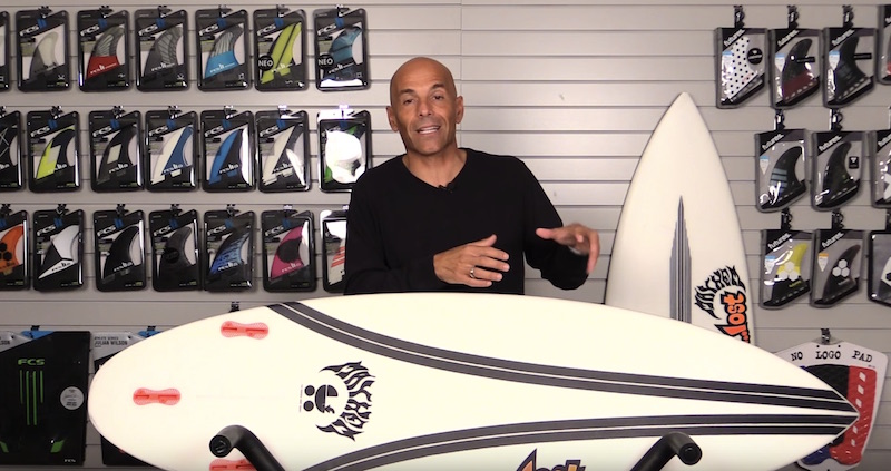 Person demonstrating a surfboard in a shop with surfboard fins displayed.