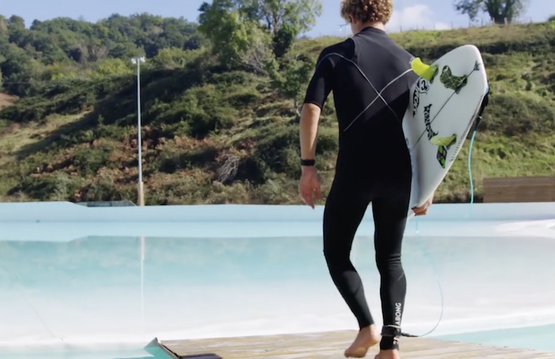 Person in wetsuit carrying a surfboard near a pool.