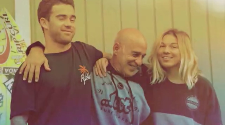 Three people smiling together, standing against a wooden wall.