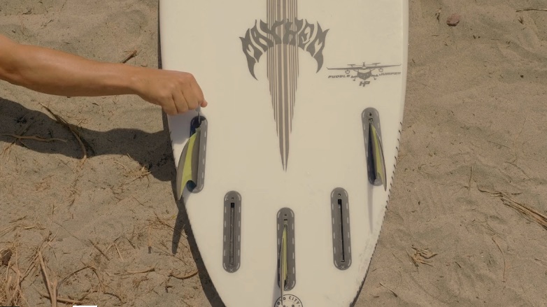 Hand adjusting fins on a surfboard resting on sand.