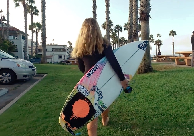 Person walking on grass carrying a surfboard near palm trees.