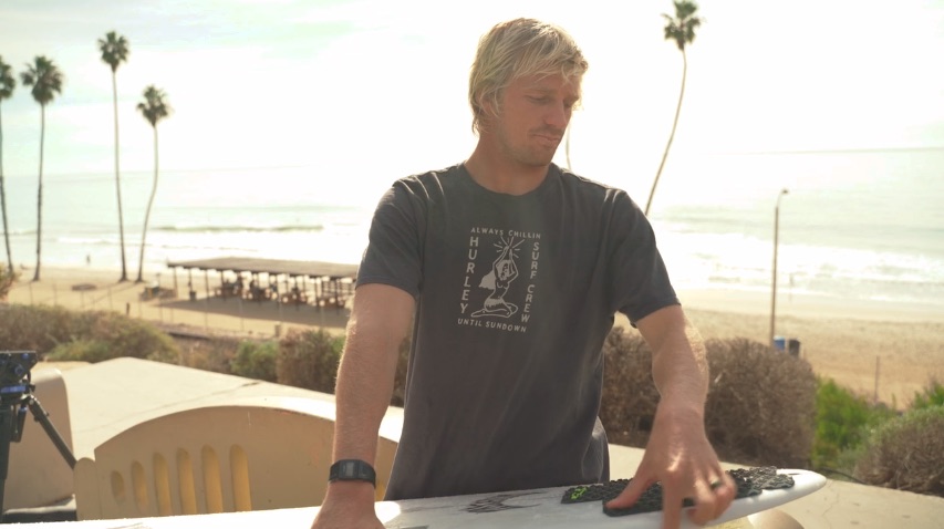 Surfer preparing his board with ocean and palm trees in background.