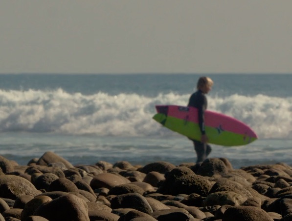 Surfer walking on rocky beach with surfboard.