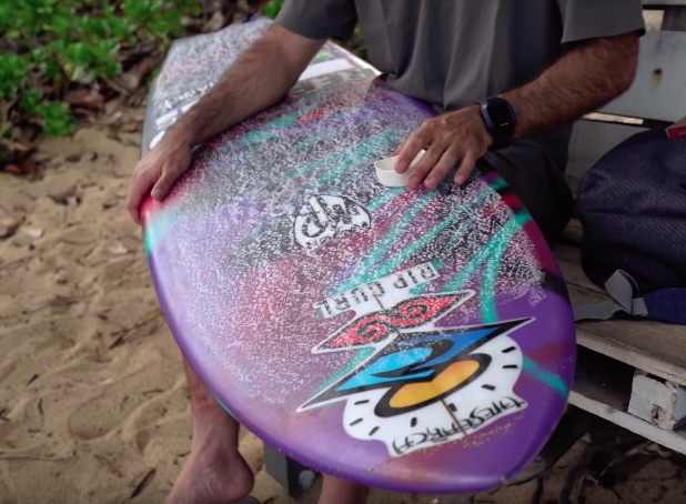 Person sanding a colorful surfboard on a beach.