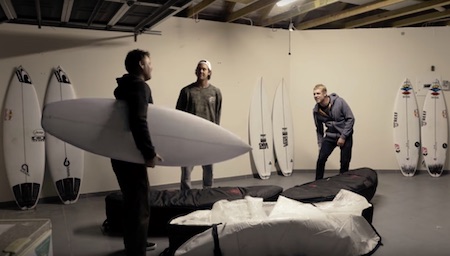 Three men in a room with surfboards and bags.