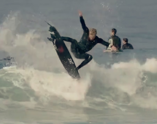 Surfer performing a trick on a wave with spectators in background.