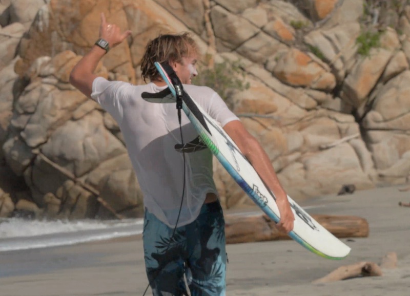 Surfer walking on the beach, holding a surfboard and signaling.