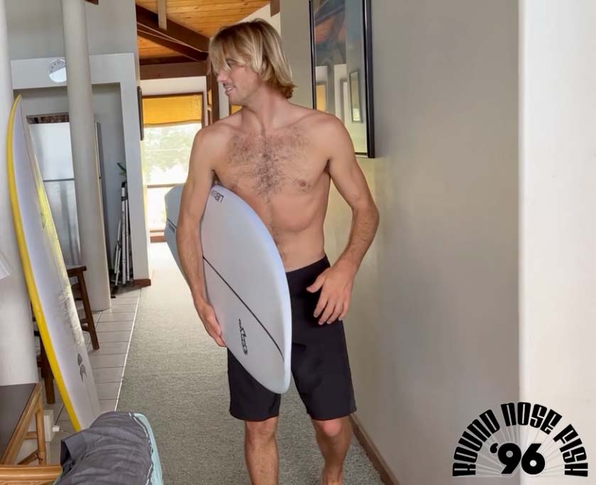 Shirtless man holding a surfboard in a hallway.