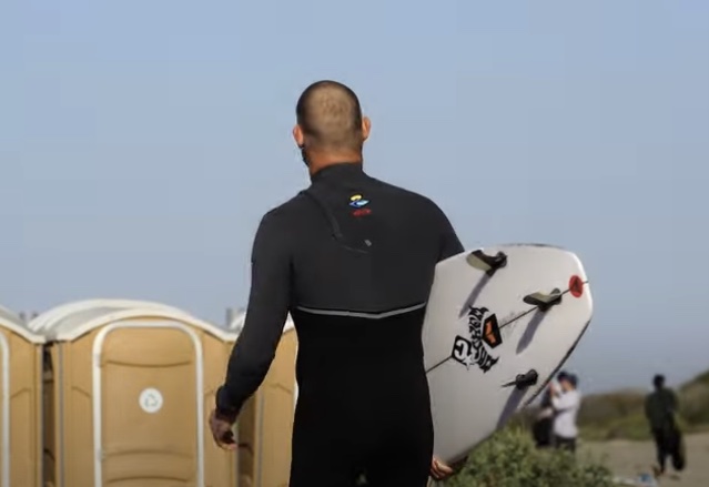 Surfer in wetsuit walking towards beach with surfboard.