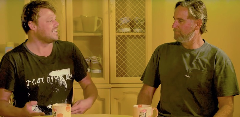 Two men sitting at a table with mugs, discussing.