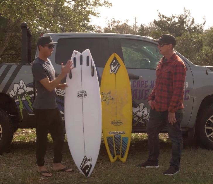 Two men discussing surfboards beside a truck in a grassy area.