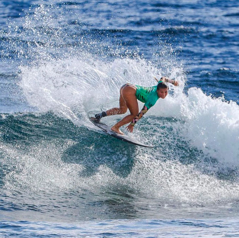 A surfer in a green top rides a wave, bending low on her board as water splashes around her. The ocean is blue and calm in the background.