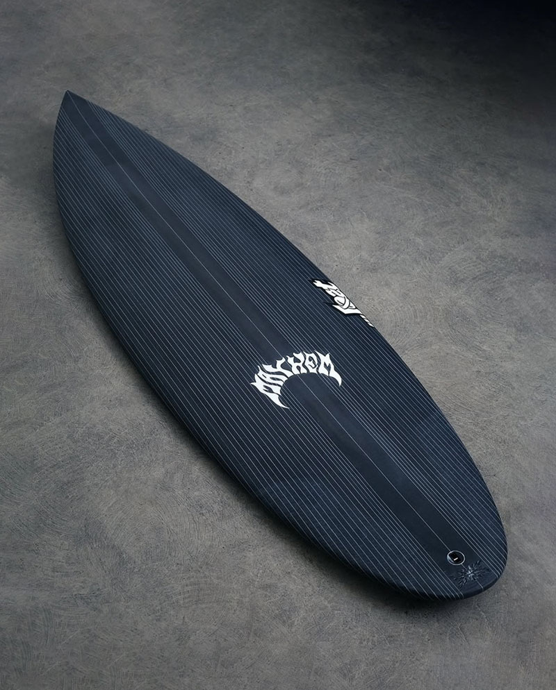 A sleek black surfboard with a striped pattern and a white logo on the side, resting on a textured gray surface. The board has a pointed nose and a single fin attachment at the tail.