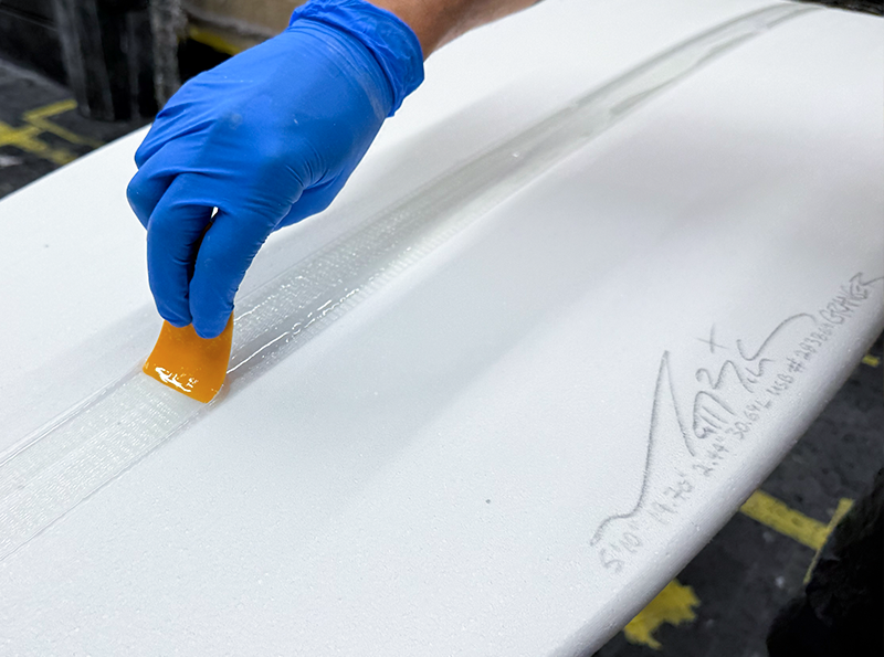 Applying adhesive to a surfboard with a gloved hand.