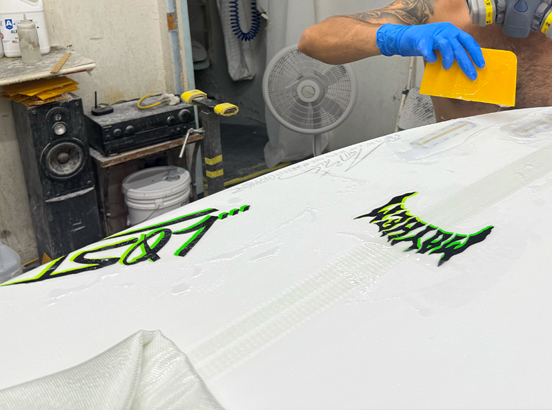 Person applying a finishing layer to a surfboard in a workshop.