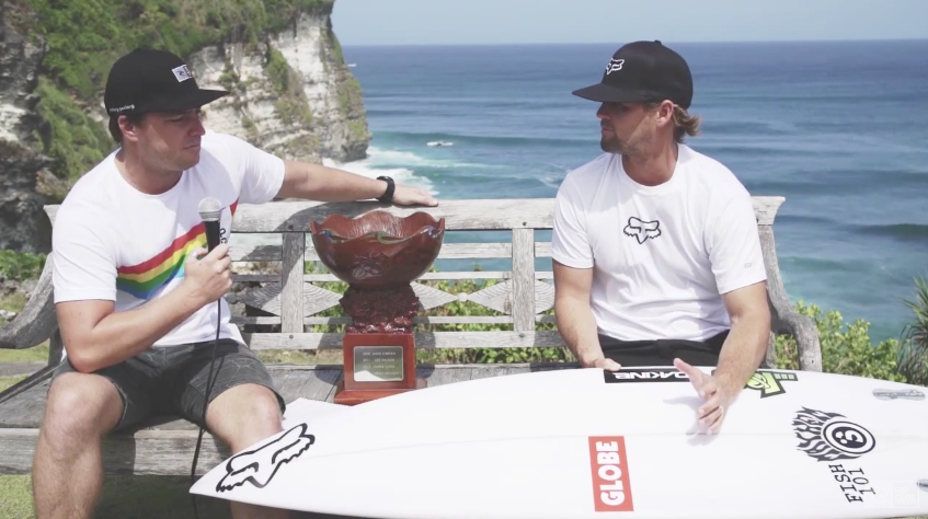 Two men sitting on a bench with a surfboard and trophy, ocean in background.