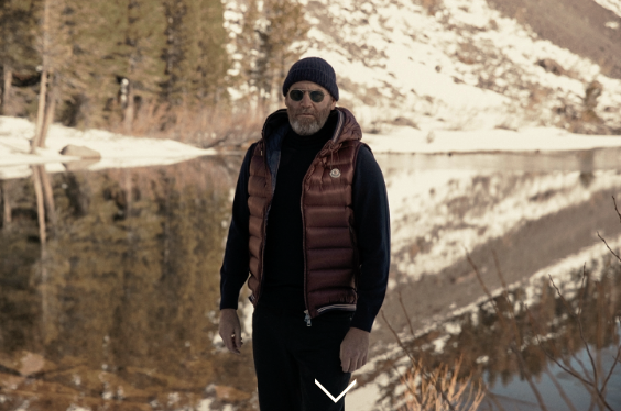 Man in a puffy vest and beanie standing by a lake.