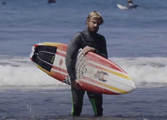 Surfer in wetsuit carrying a surfboard at the beach.