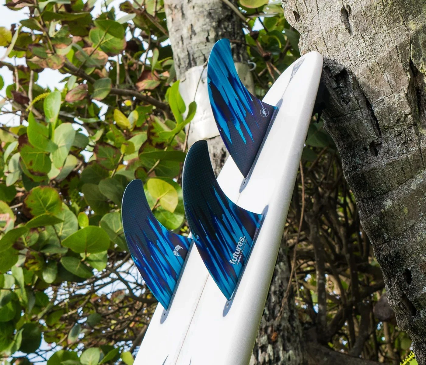 Surfboard fins mounted on a white surfboard against greenery.