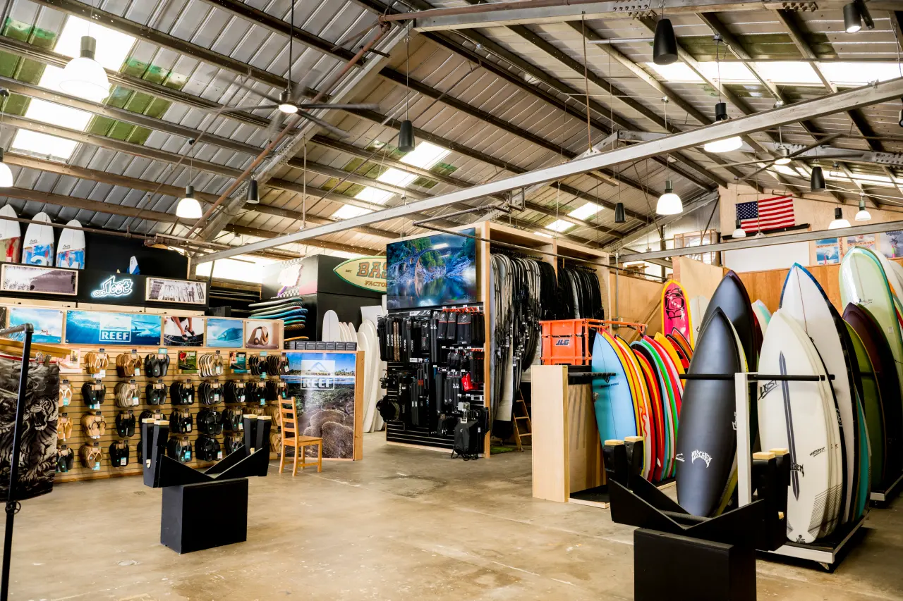 A surf shop interior featuring various surfboards displayed on racks, along with flip-flops and accessories on shelves. The space has a high ceiling with lights and an American flag visible in the background.